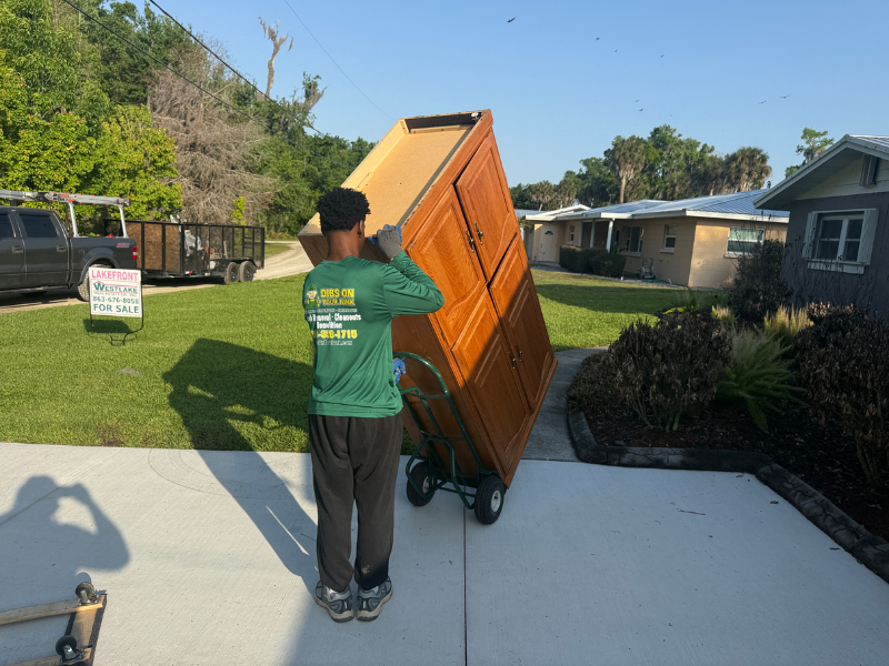 Dibs On Your Junk crew member in branded green shirt hauling large wooden armoire on hand truck during furniture removal job in Davenport, FL